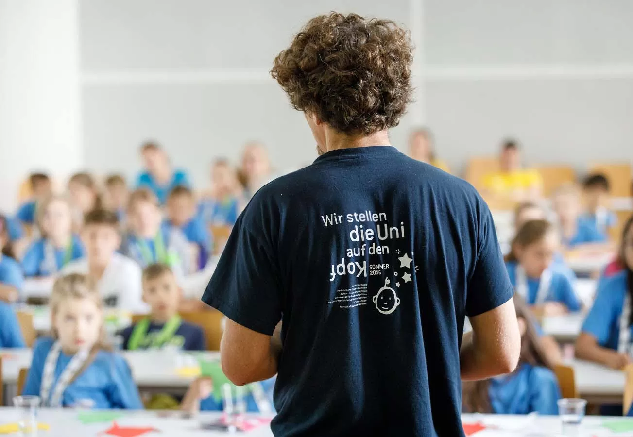 ein Lehrender trägt das KinderuniWien T-Shirt mit dem Motto "Wir stellen die Uni auf den Kopf!"